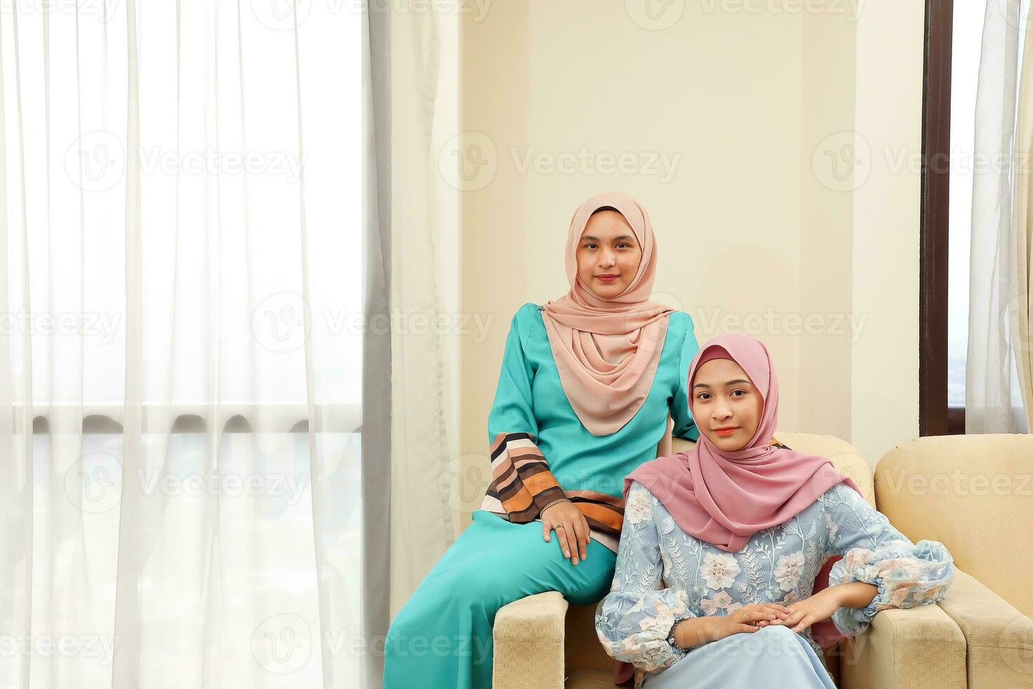 Two young Asian Malay Muslim woman wearing headscarf at home office student sitting on sofa look at camera photo
