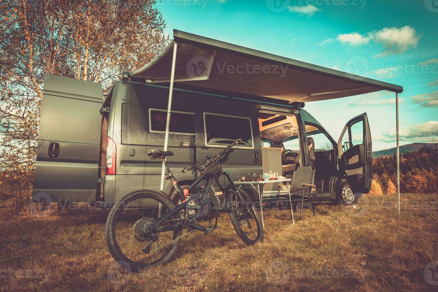 Family Motorhome Trip for an Active Weekend Away. Mountain Bike Stands Under the Tent in Front of the Camper. Light in the Van is On. Table, Chairs and Chessboard are Prepared for a Chess Play. Beautiful Autumn Vibes, Mountainous and Wooded Spot. photo