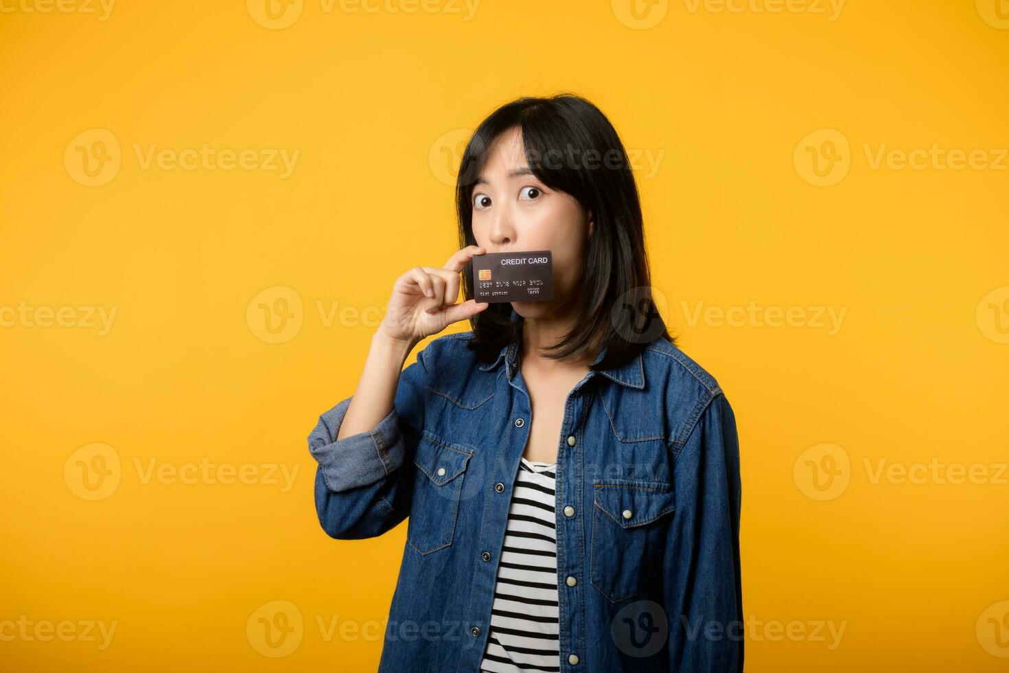 retrato hermosa joven asiático mujer contento sonrisa vestido en mezclilla chaqueta demostración teléfono inteligente y crédito tarjeta aislar en amarillo estudio antecedentes. compras en línea teléfono inteligente solicitud concepto foto