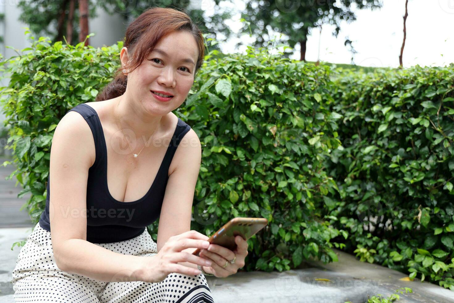 beautiful attractive middle age southeast asian woman sitting relaxing reading texting smart phone in urban green garden background photo