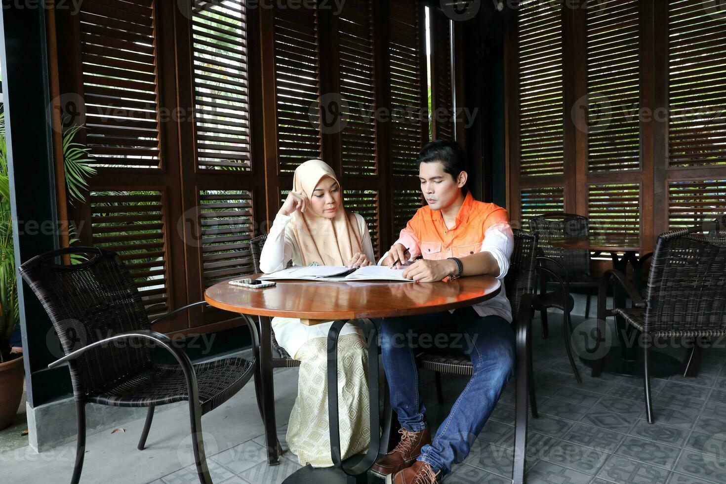 Young asian malay man woman with headscarf sit at rustic cafe table talk discuss business study read photo