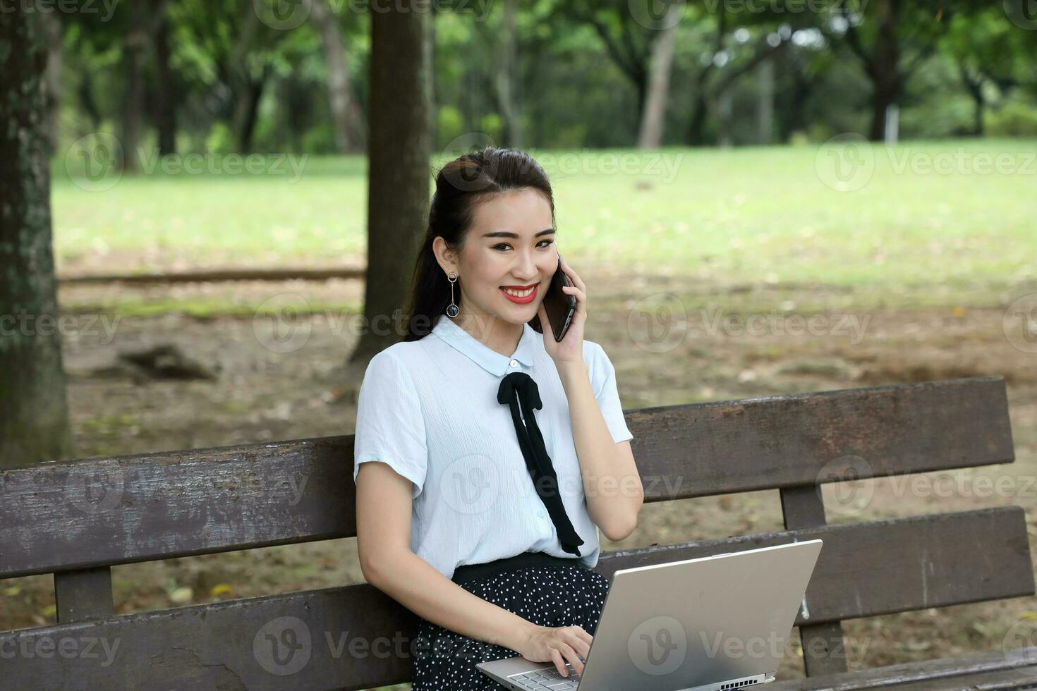 Young asian chinese woman outdoor on park bench use laptop computer think read look happy wonder smile front talk on phone photo