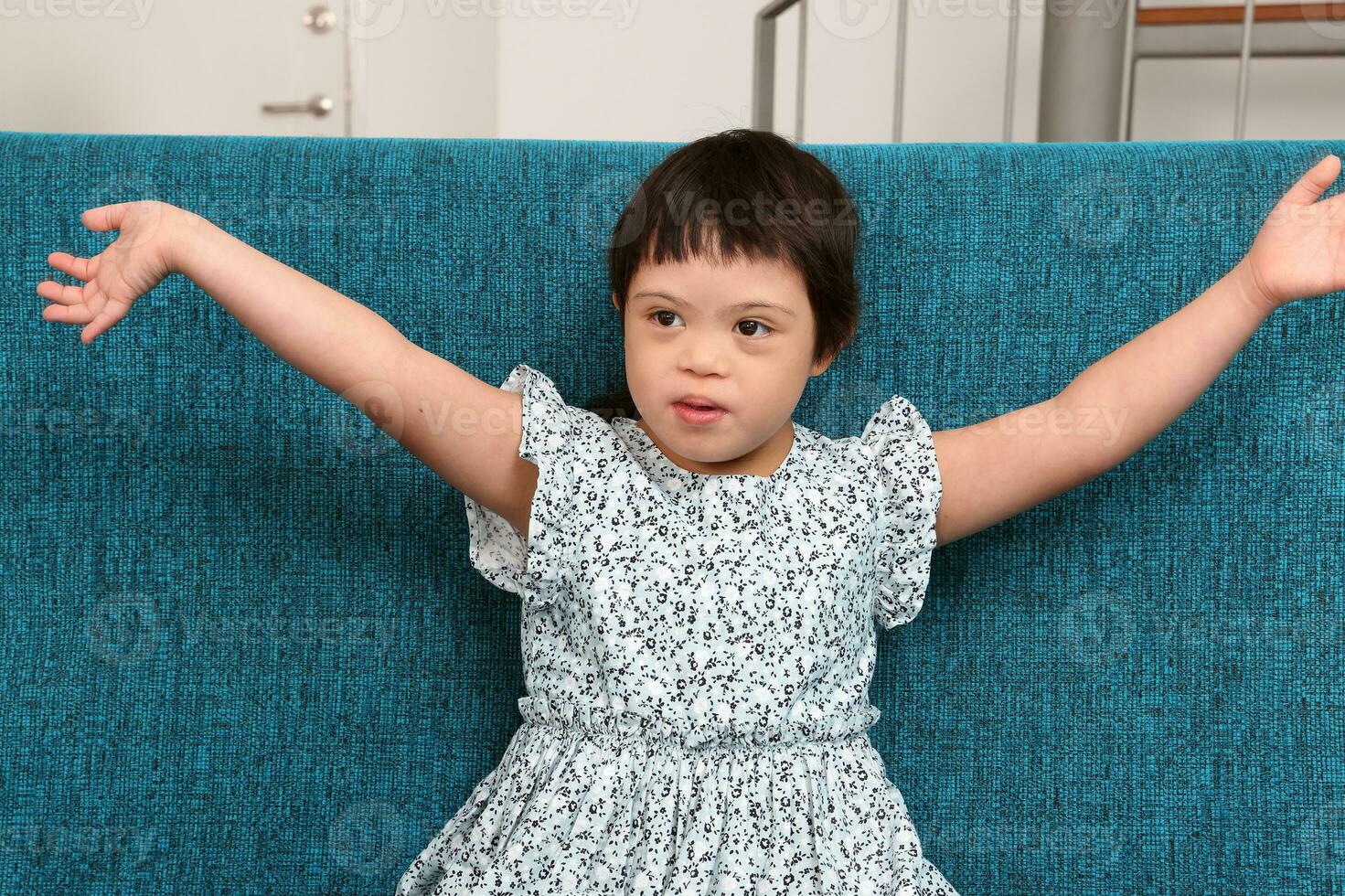 Southeast Asian small little girl child sitting on blue sofa look happy smile pose open arm happy hug sing. She have down syndrome photo