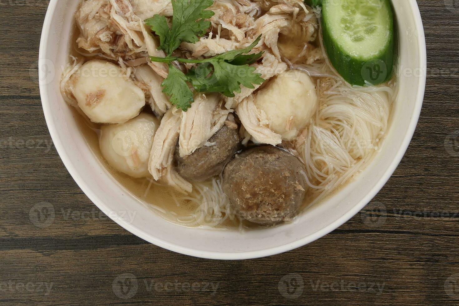 Clear white rice vermicelli beehoon noodle soup with chicken beef meat ball shredded cucumber in white bowl chopsticks spoon chili soya sauce parsley garnish photo