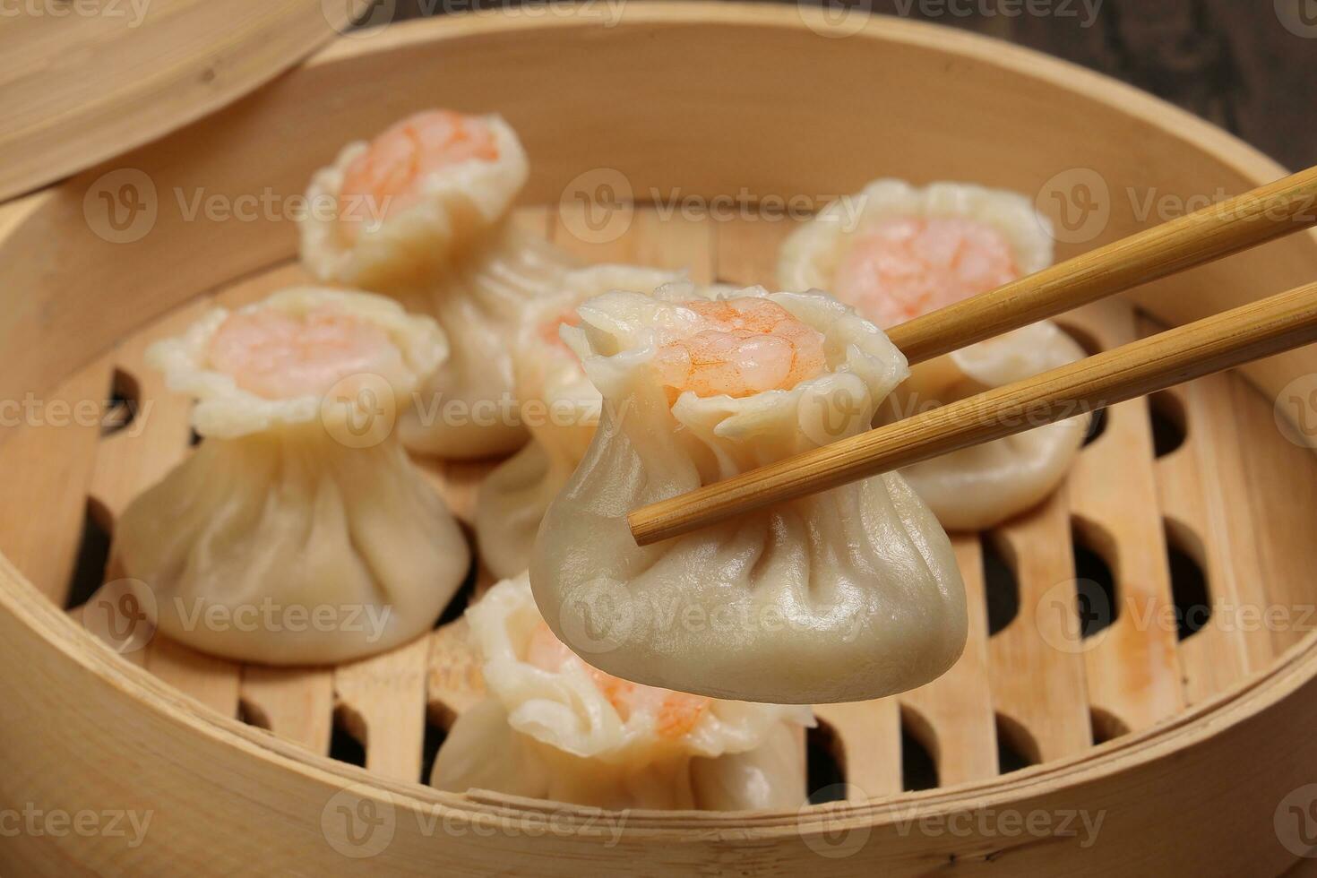 Prawn shrimp shaomai dim sum dumpling picked up by chopsticks in bamboo steamer on rustic wood background photo
