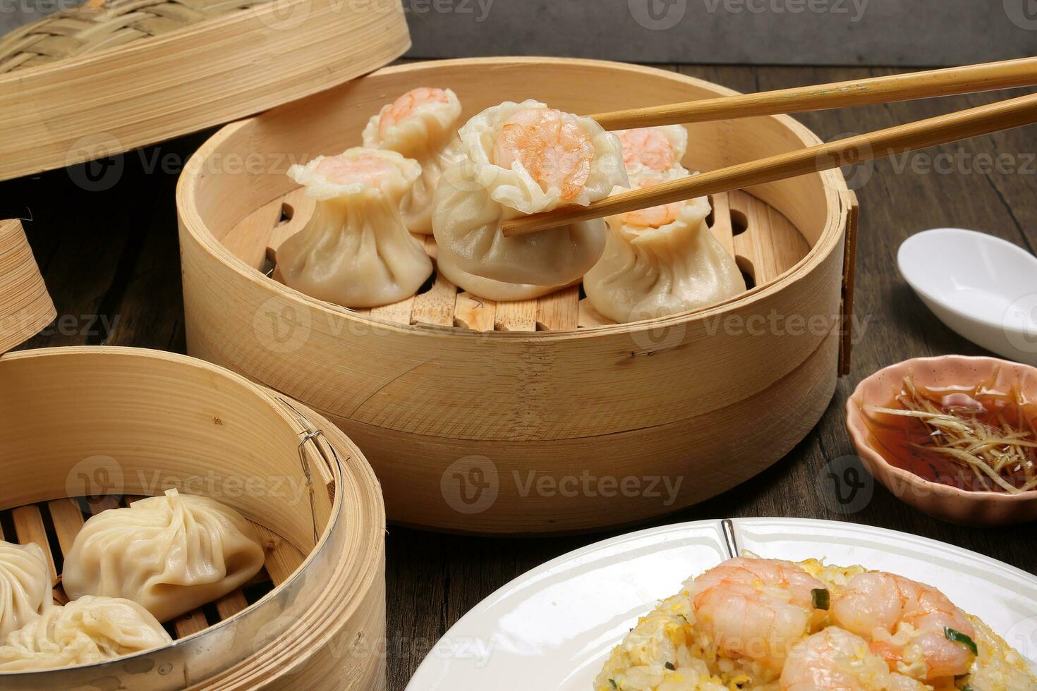 Prawn shrimp shaomai pickup by chopsticks Xiao long bao dim sum dumpling chicken prawn fish seafood vegetable in bamboo steamer fried rice on plate sauce soup spoon over rustic background photo