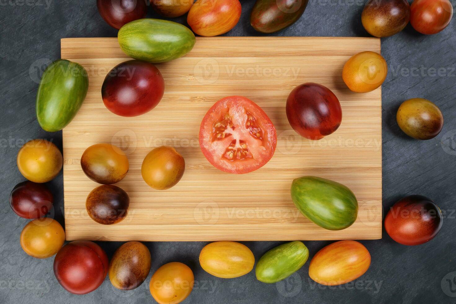 Red orange yellow green tomato mix variety on around wooden chopping board frame copy text space over black slate stone background photo