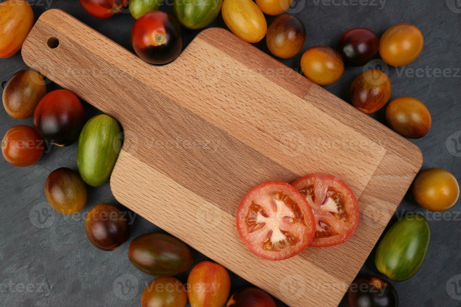 Red orange yellow green tomato mix variety on around wooden chopping board frame copy text space over black slate stone background photo