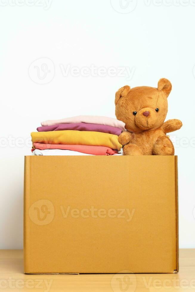 Kid Donation, Charity, Volunteer, Giving and Delivery Concept. Hand donate Bear doll and Clothes into cardboard box at home for support and help poor, refugee and homeless people. Copy space for text photo