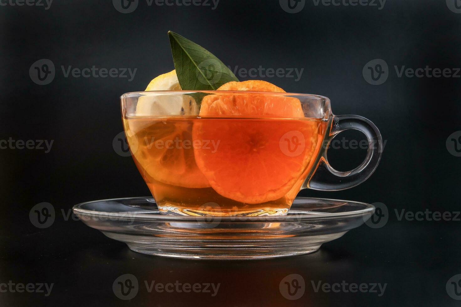 Liquid tea lemon orange slice green leaf cinnamon stick in transparent glass teacup saucer on black background photo