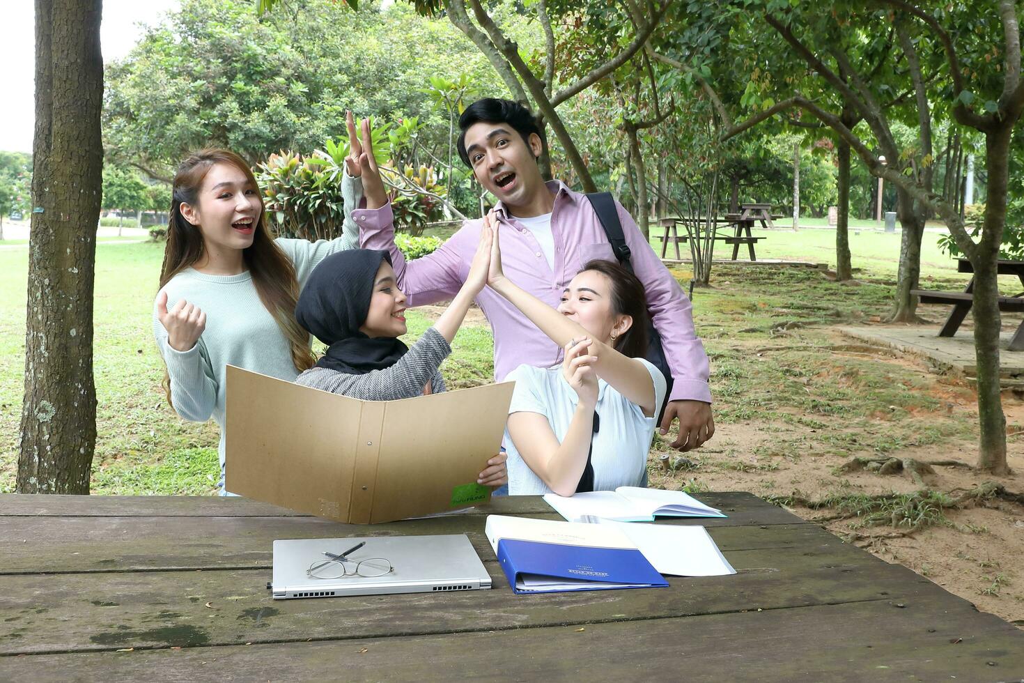 Young asian malay chinese man woman outdoor on park bench table study talk discuss point laptop file book happy high five photo