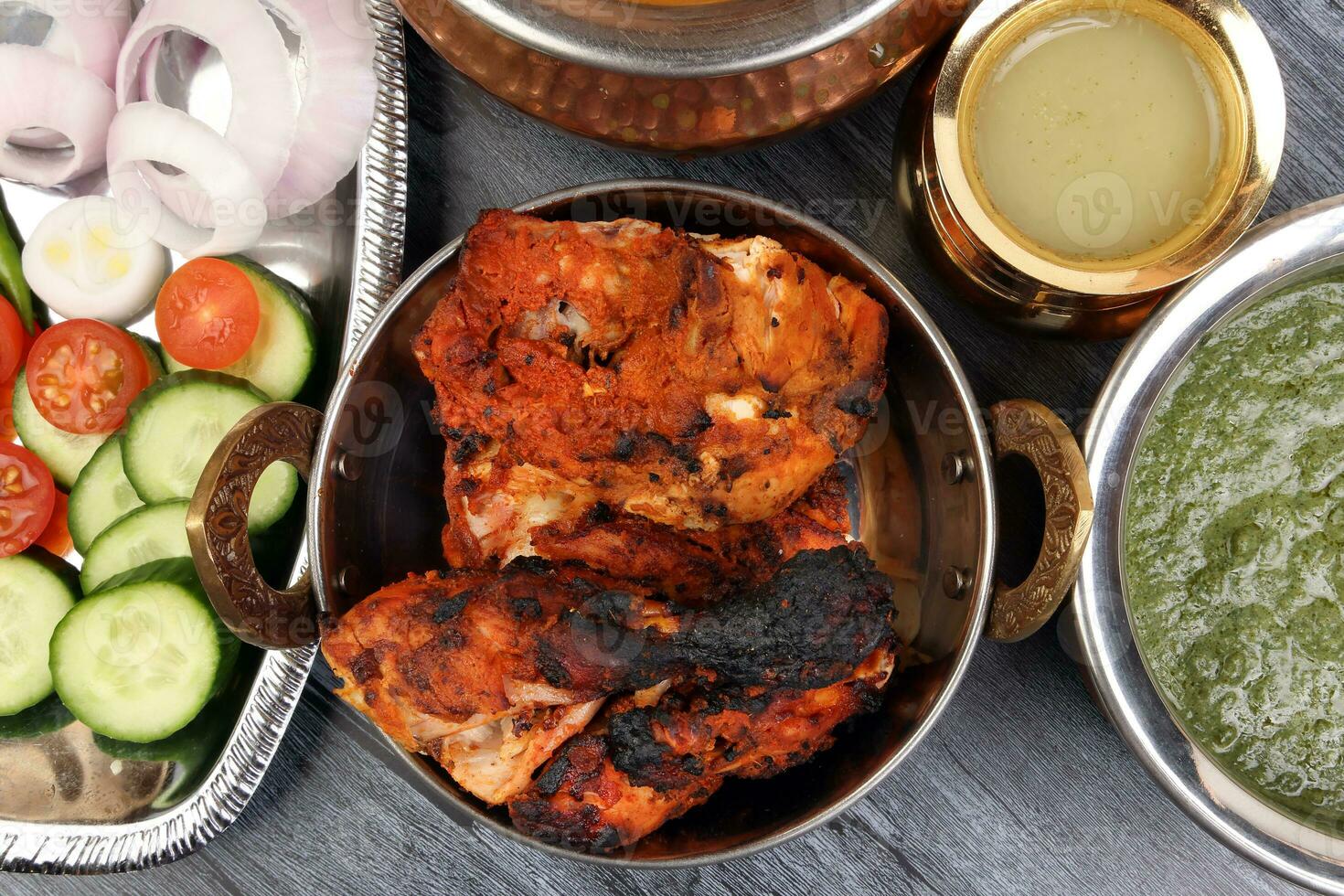 Spicy red Chicken grilled tikka tandoori nan bread green spinach curry yogurt sauce dal tomato cucumber onion salad set in metal stainless steel brass copper pot on black char wood background photo
