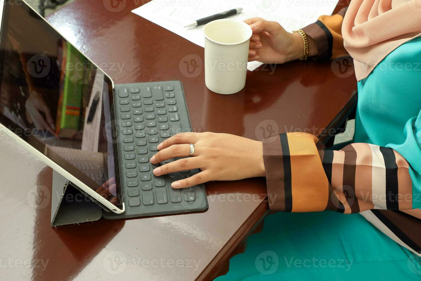 young Asian Malay Muslim woman at home office student sitting at table computer pone book document study type hand hold coffee tea cup photo