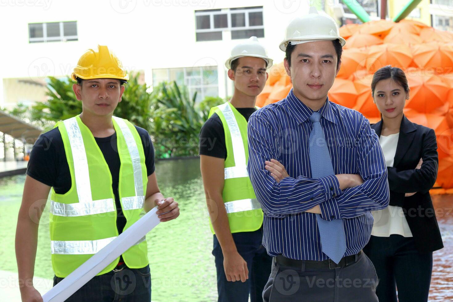 South East Asian young Malay Chinese man woman safety helmet vest formalwear stand at construction site blueprint plan clipboard look at camera photo