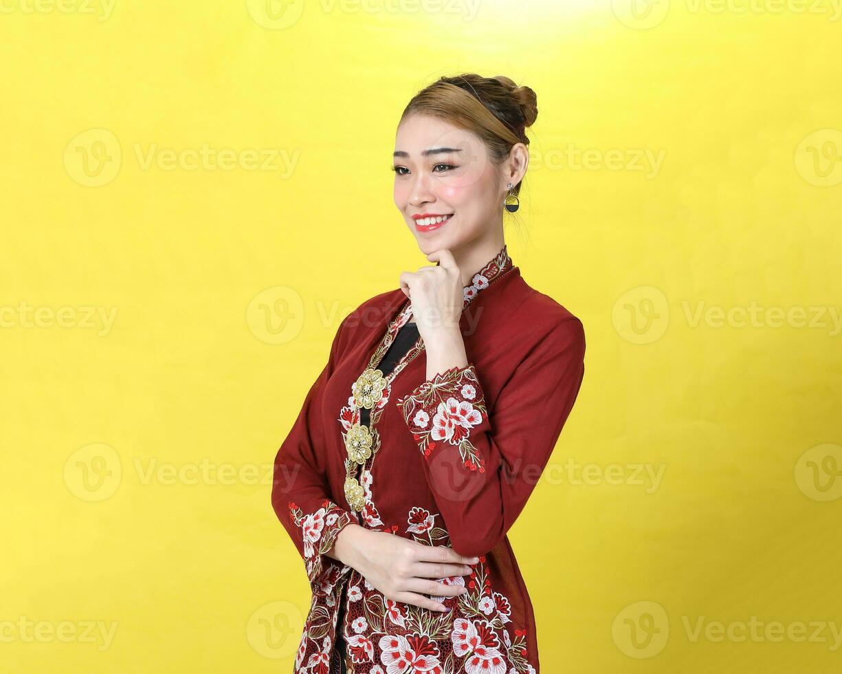 Asian woman traditional red maroon kebaya sharong on yellow paper background posing stand happy finger on chin photo