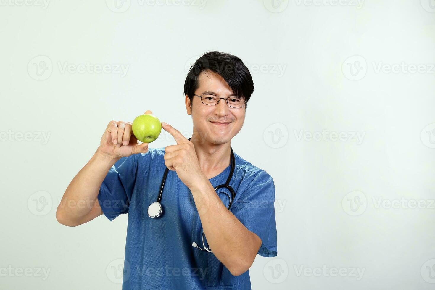 Young Asian male doctor wearing apron uniform tunic stethoscope holding pointing showing eating healthy green apple photo