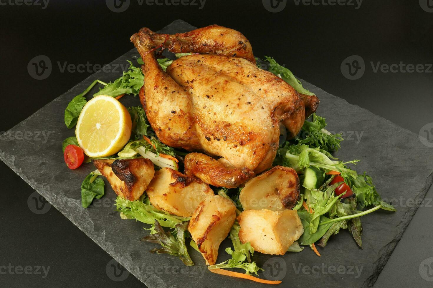 Whole roasted grilled chicken poultry bird with baked potato vegetable salad tomato lemon on black slate stone cutting board black background photo