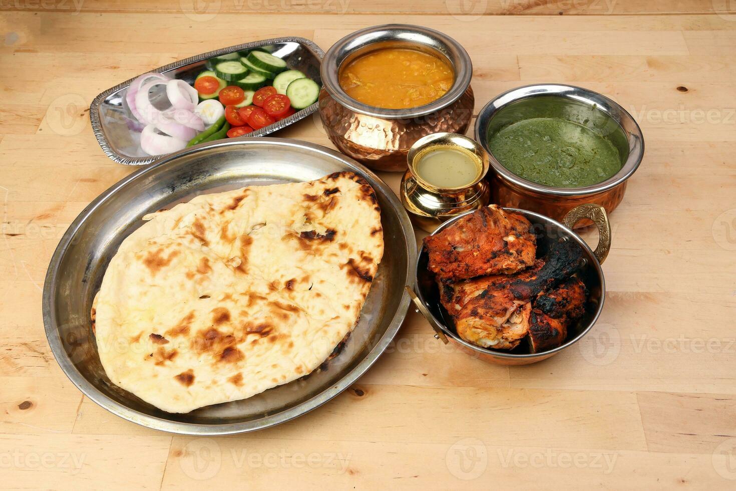Spicy red Chicken grilled tikka tandoori nan bread green spinach curry yogurt sauce dal tomato cucumber onion salad set in metal stainless steel brass copper pot on light wood background photo