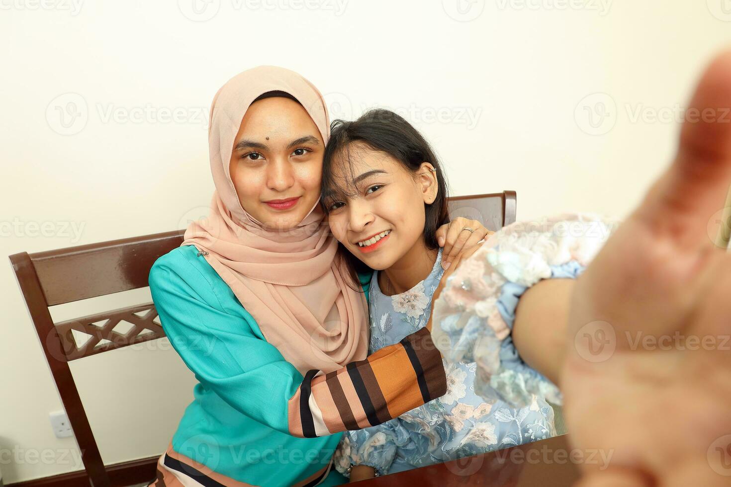 Two young Asian Malay Muslim woman wearing headscarf at home office student sitting at table phone computer book document selfie self portrait hug care affection POV point of view photo