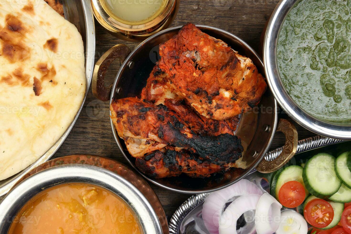 Spicy red Chicken grilled tikka tandoori nan bread green spinach curry yogurt sauce dal tomato cucumber onion salad set in metal stainless steel brass copper pot on rustic vintage wood background photo