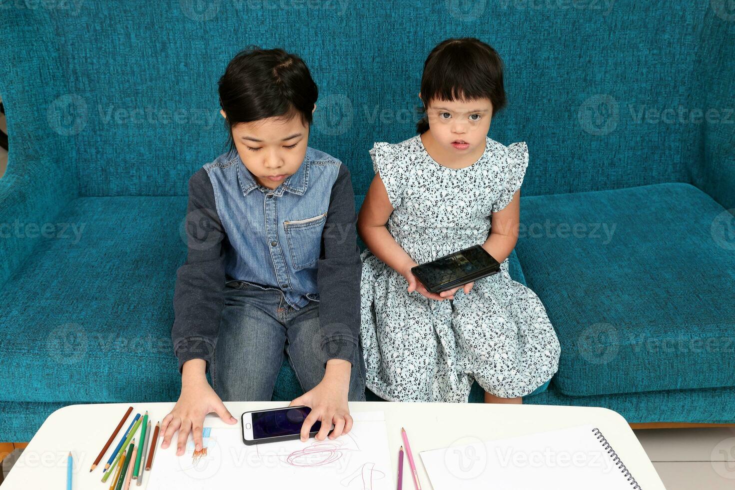Two little south east Asian girl child malay Chinese study play draw use smart phone on blue fabric sofa they are autistic and down syndrome photo