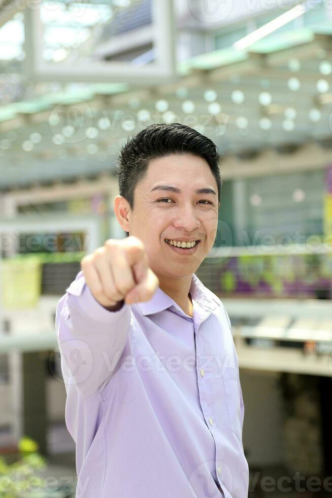 Young south east Asian man business office outdoor stand look at camera point finger happy photo