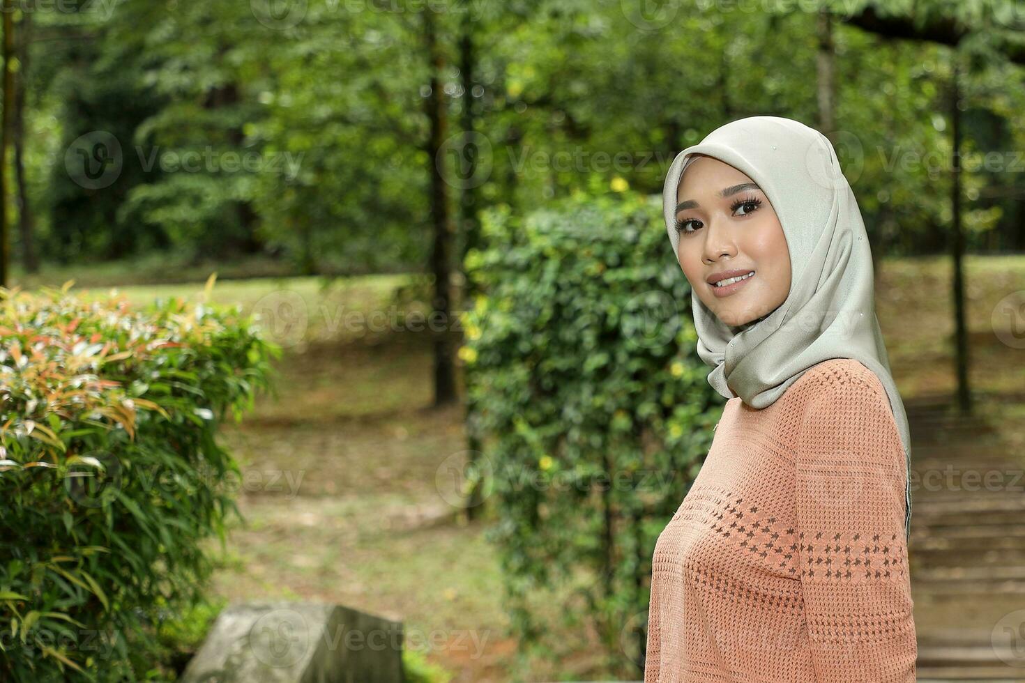 One Asian Malay Muslim woman in headscarf posing smile look forward towards camera confident outdoor green park photo