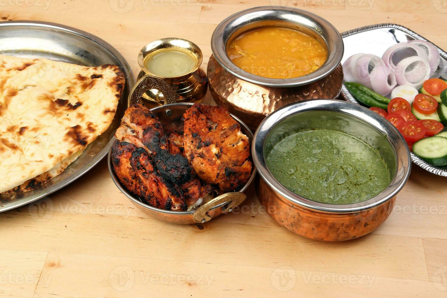 Spicy red Chicken grilled tikka tandoori nan bread green spinach curry yogurt sauce dal tomato cucumber onion salad set in metal stainless steel brass copper pot on light wood background photo