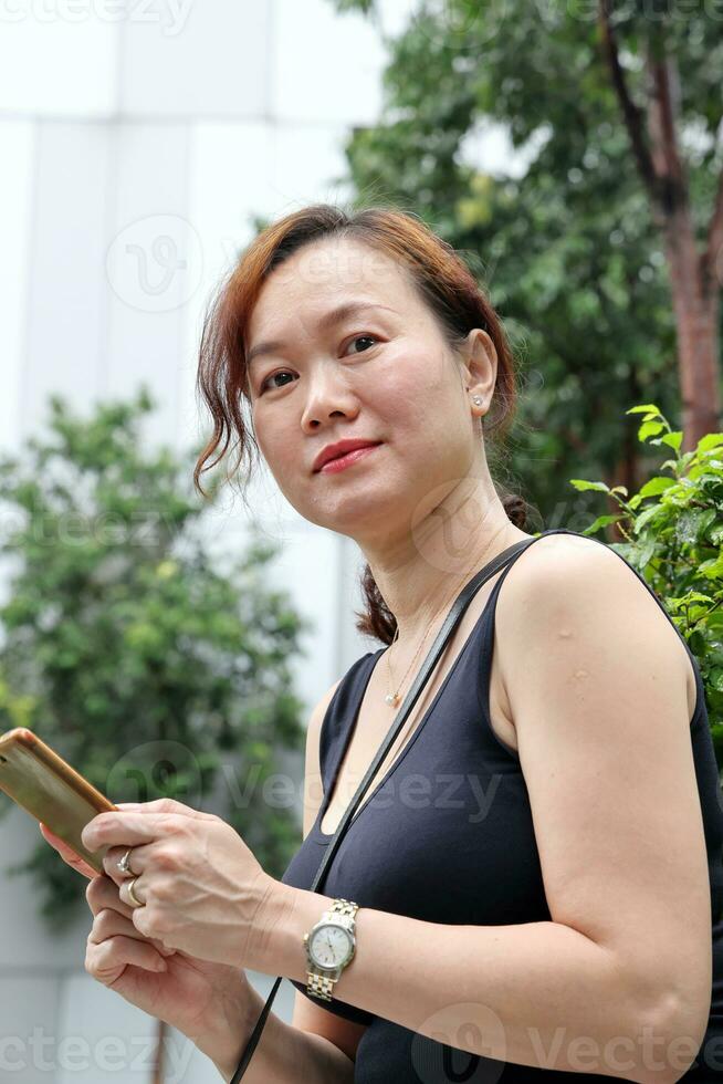 beautiful attractive middle age southeast asian woman sitting relaxing reading texting smart phone in urban green garden background photo