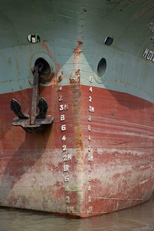 Large ship front close up shot on a riverbank. Rusty anchor hanging on the ship. Old ship front with scratches and water meter. Cargo ship docked on the riverbank. Rusty marine life ship front shot. photo