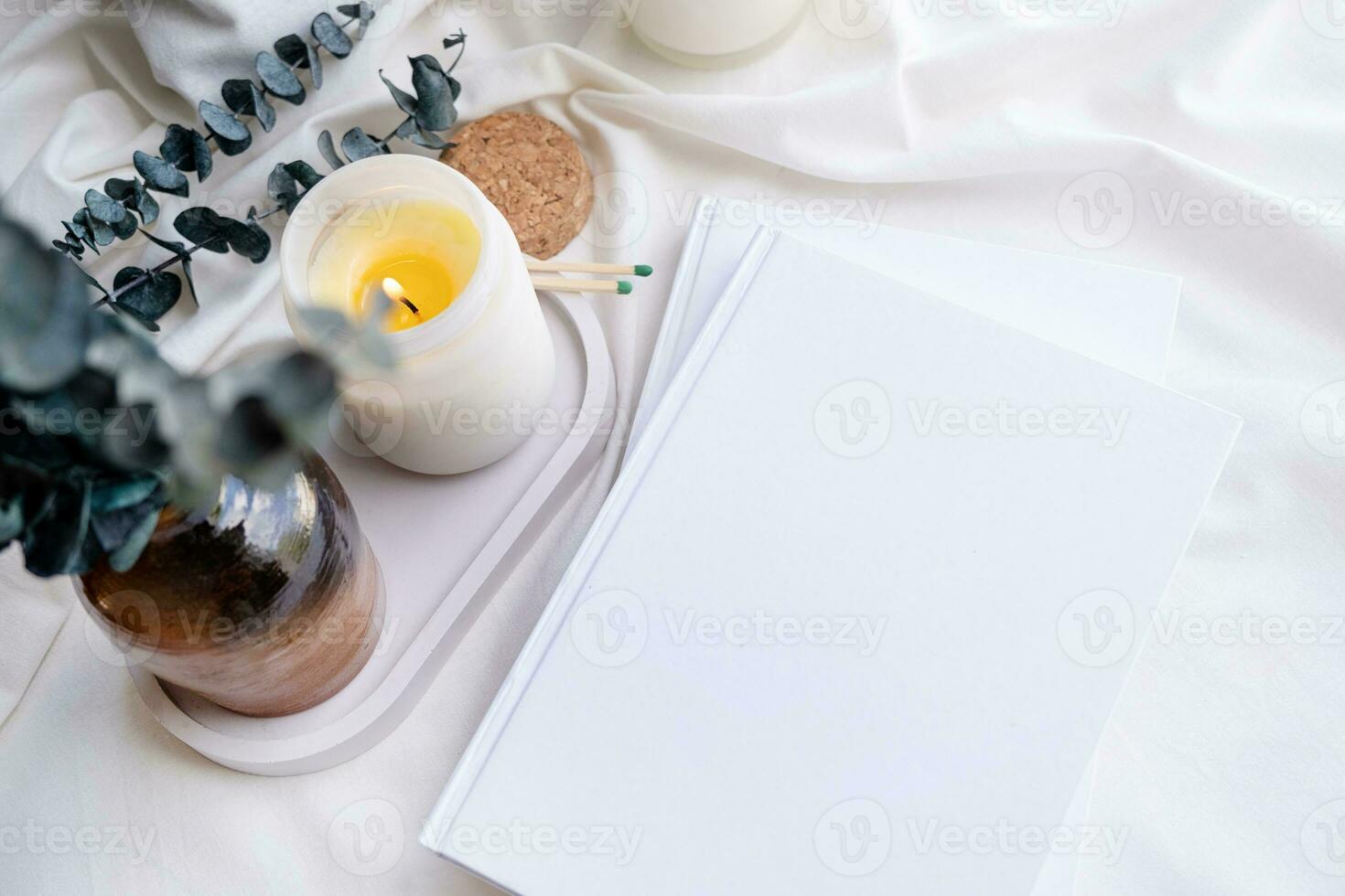 Soy wax aroma candle in white jar on bed with eucalyptus leaves. Candle and book mockup design photo