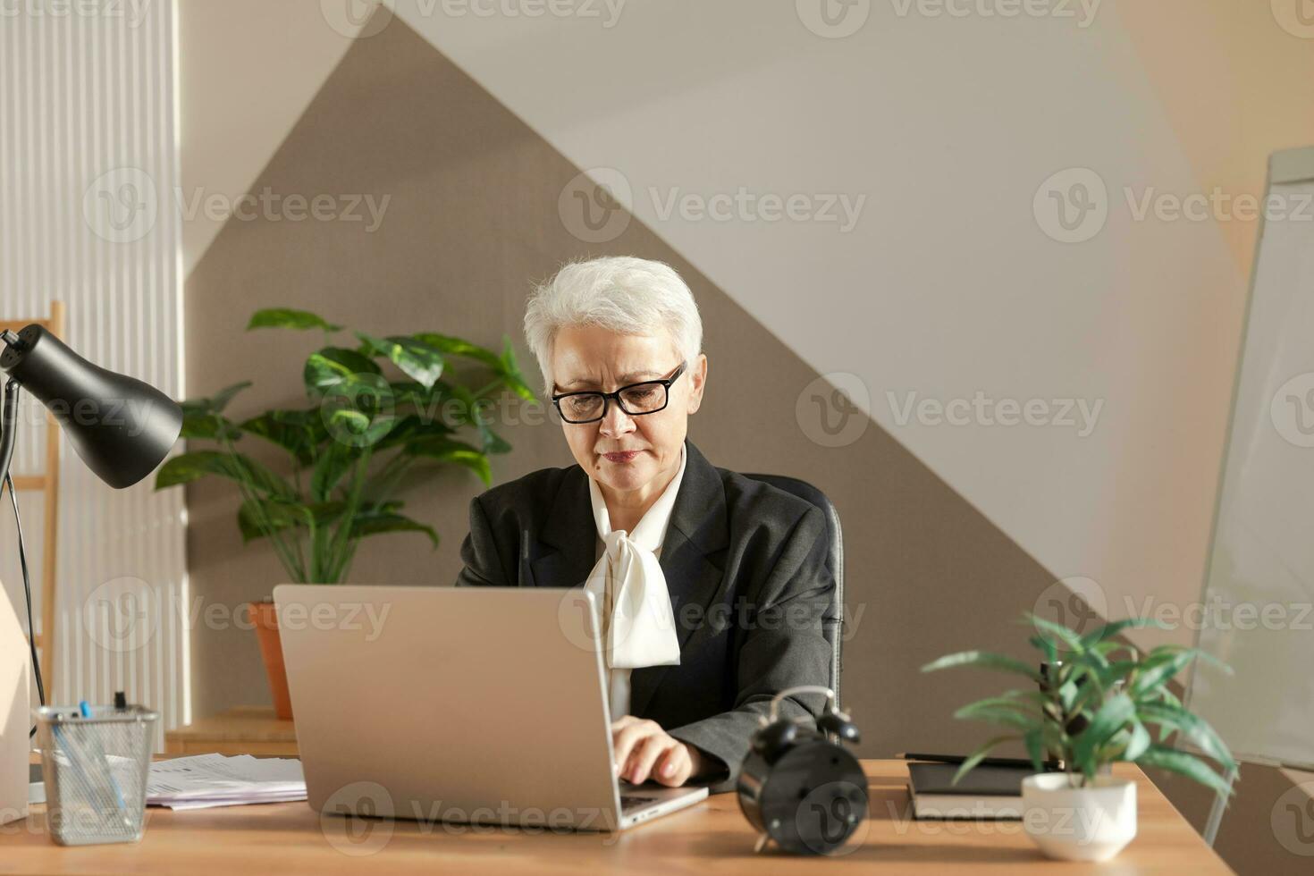 Confident stylish european middle aged senior woman using laptop at workplace. Stylish older mature 60s gray haired lady businesswoman sitting at office table. Boss leader teacher professional worker. photo