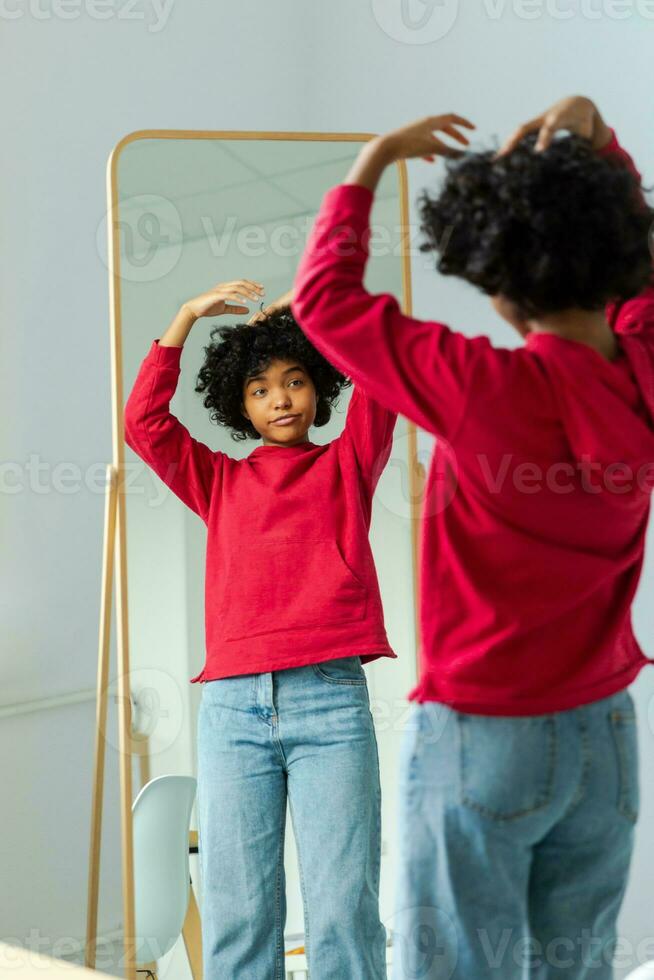 Love yourself. Beautiful young smiling african american woman dancing touching curly hair enjoying her mirror reflection. Black lady looking at mirror looking confident and happy. Self love concept. photo