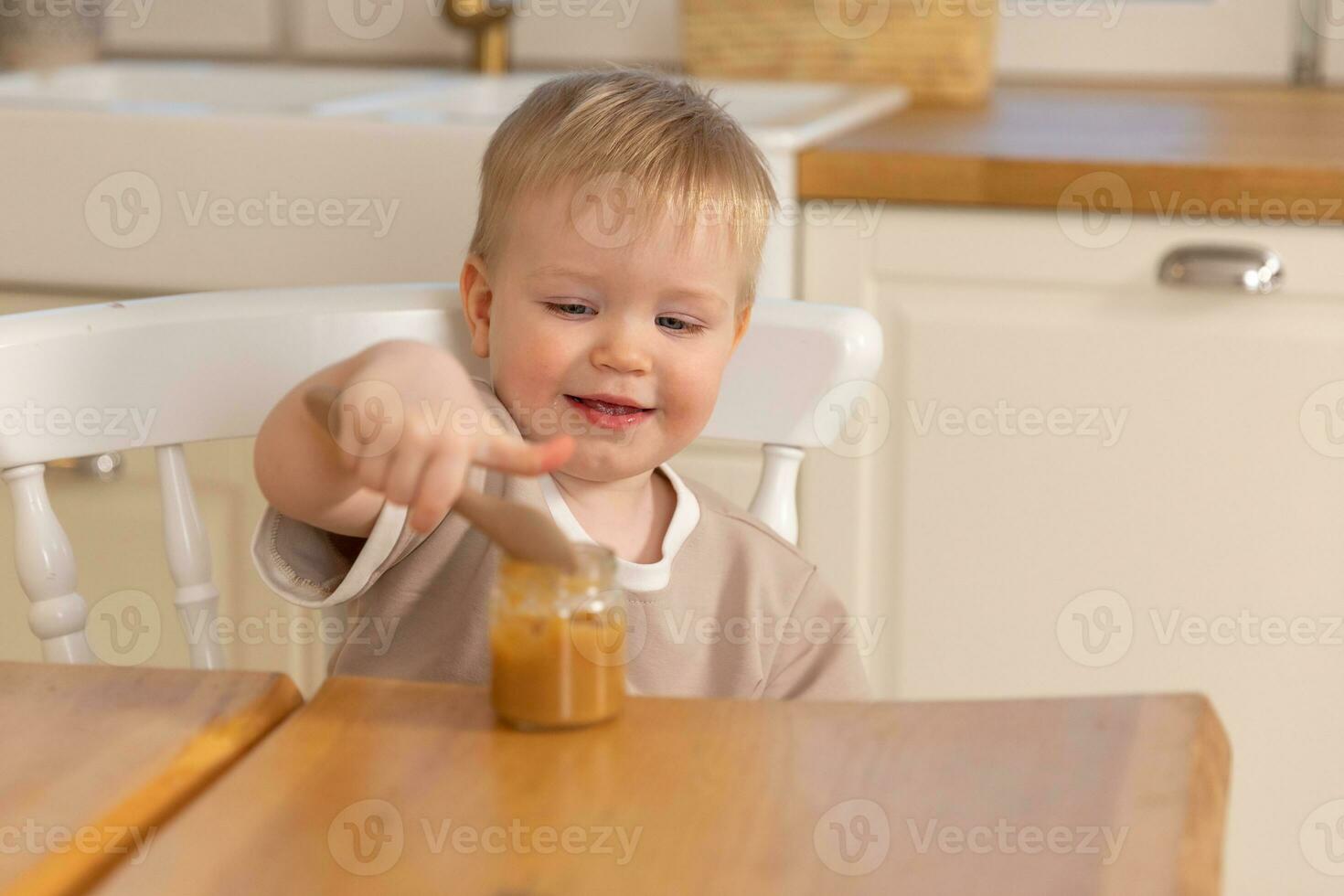 Happy family at home. Baby boy feeding himself in kitchen. Little boy with messy funny face eats healthy food. Child learns eat by himself holding spoon. Self feeding. photo