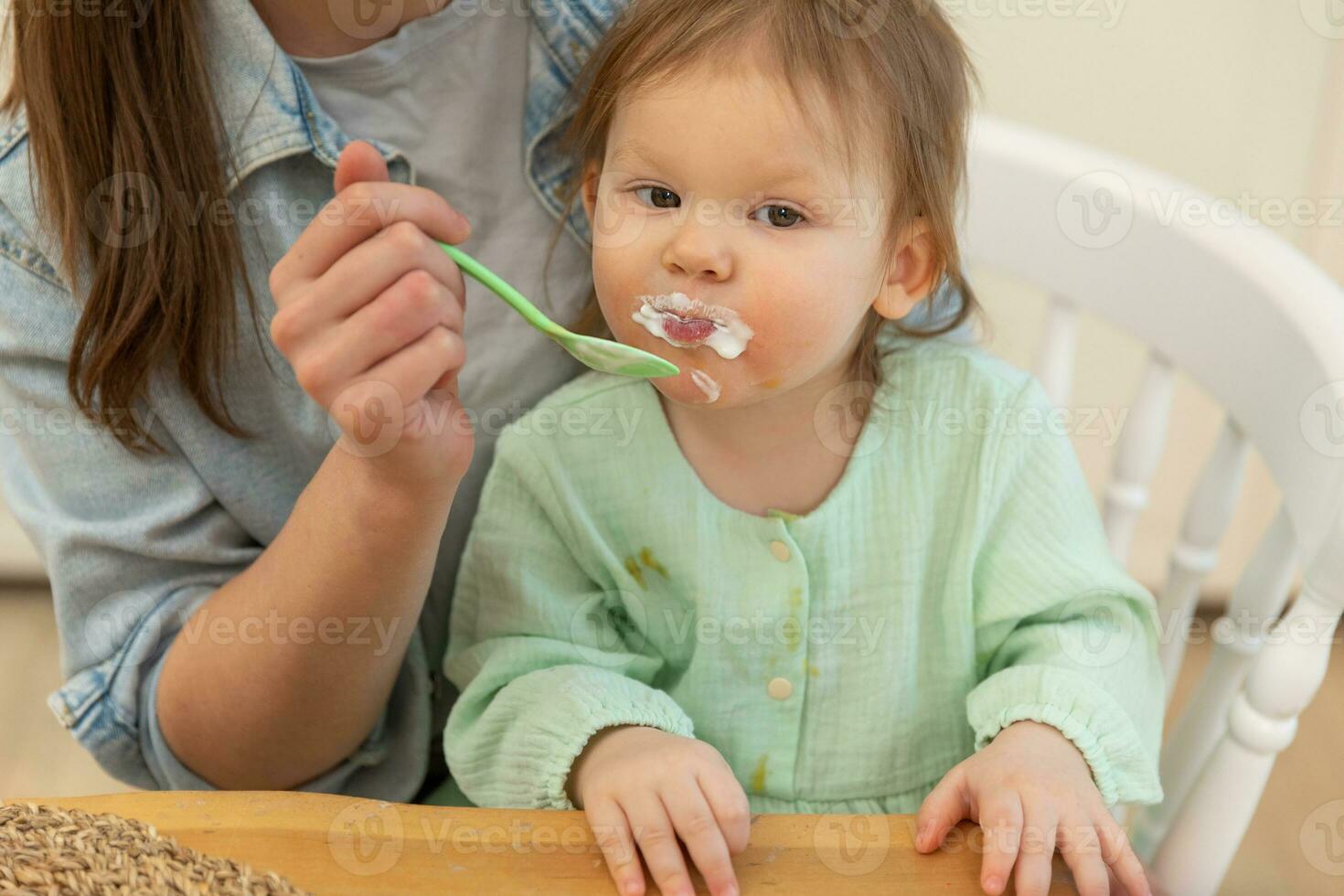 Happy family at home. Mother feeding her baby girl from spoon in kitchen. Little toddler child with messy funny face eats healthy food at home. Young woman mom giving food to kid daughter. photo