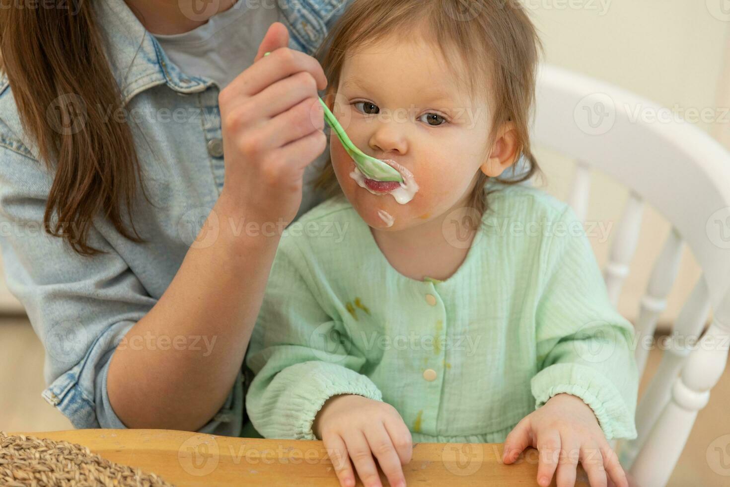 Happy family at home. Mother feeding her baby girl from spoon in kitchen. Little toddler child with messy funny face eats healthy food at home. Young woman mom giving food to kid daughter. photo