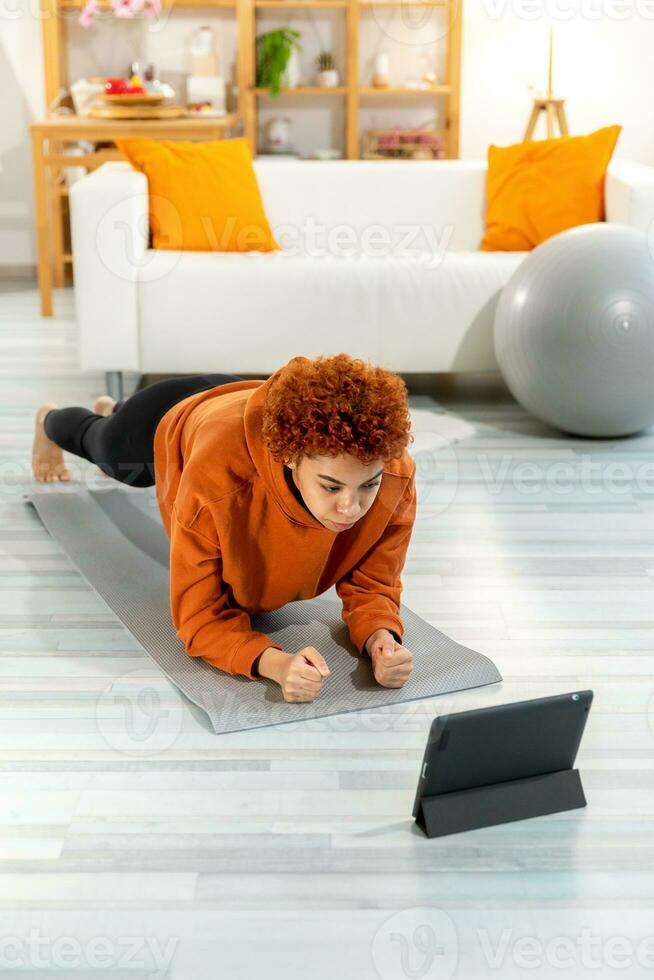 aptitud rutina de ejercicio capacitación. joven sano ajuste africano niña haciendo tablón ejercicio en yoga estera en piso a hogar. atlético mujer en ropa de deporte formación pilates deporte y aptitud física. foto