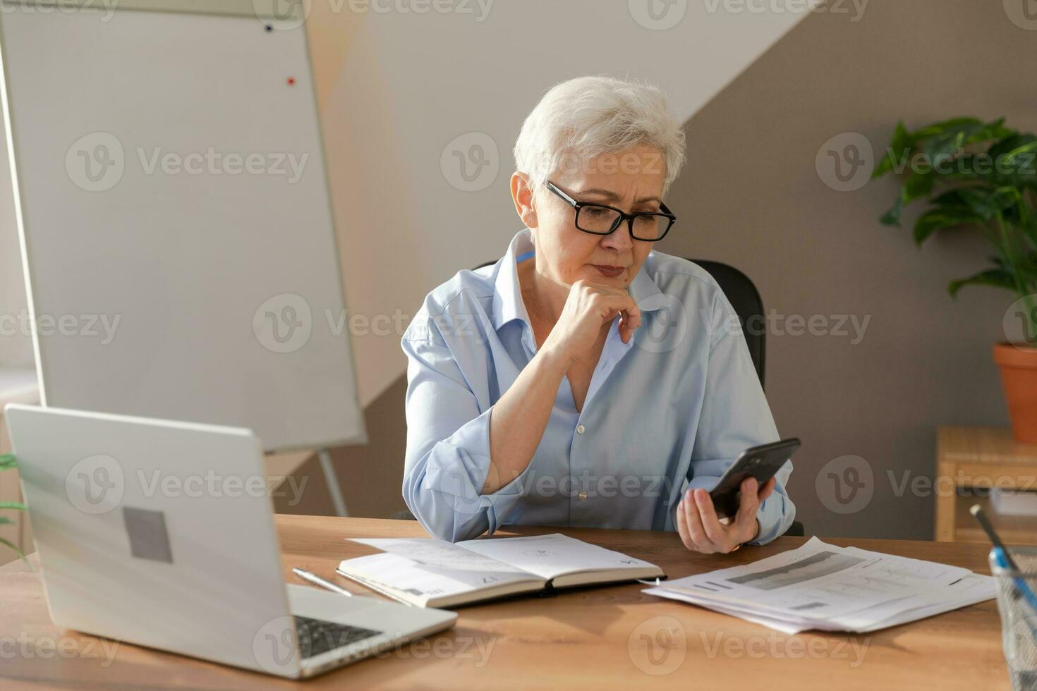 Confident stylish european middle aged senior woman using smartphone at workplace. Stylish older mature 60s gray haired lady businesswoman with cell phone in office. Boss leader using internet apps. photo