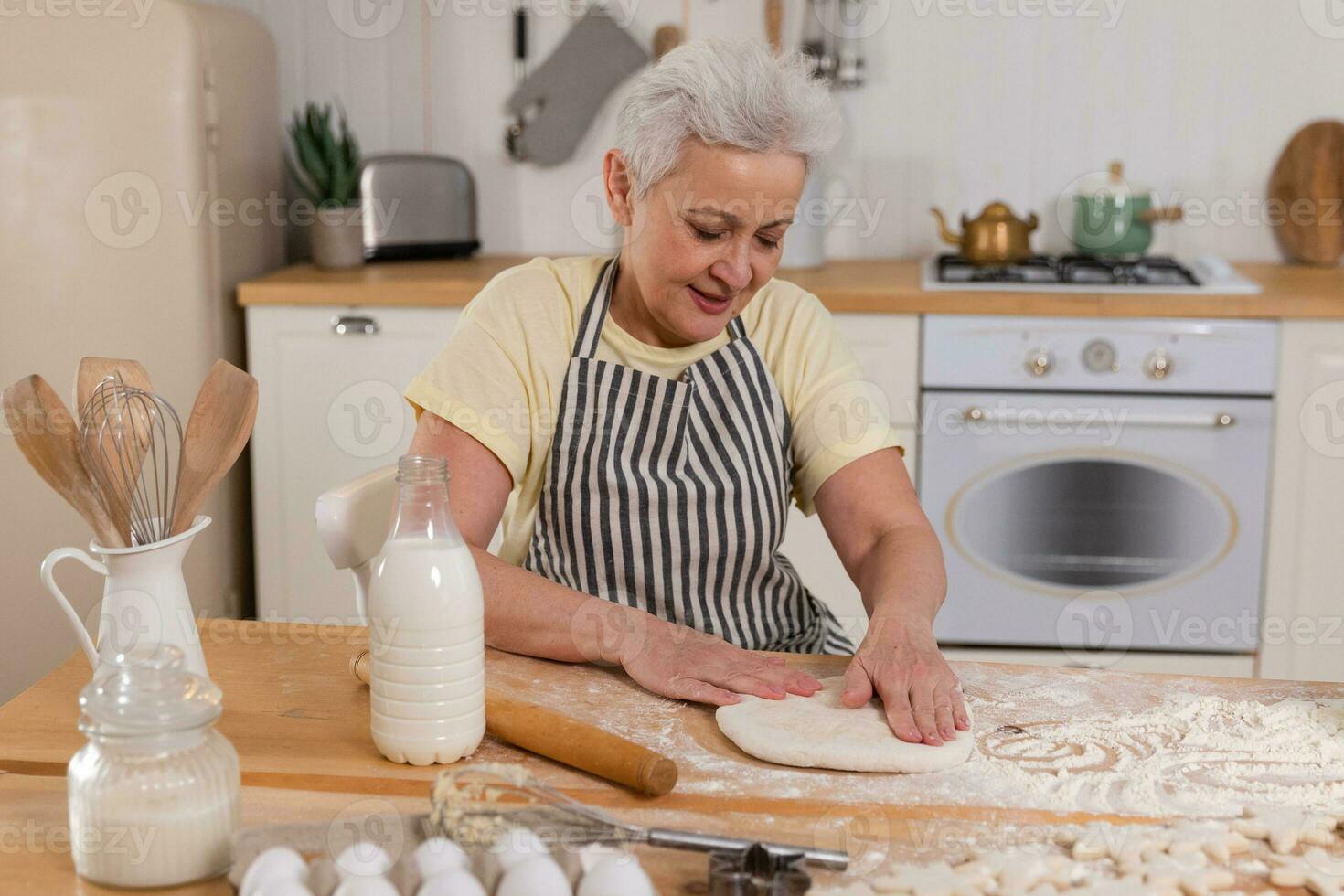 Happy senior woman cooking in kitchen. Stylish older mature gray haired lady grandmother knead dough bake cookies. Old grandma cook homemade food. Household housewife housework concept. photo