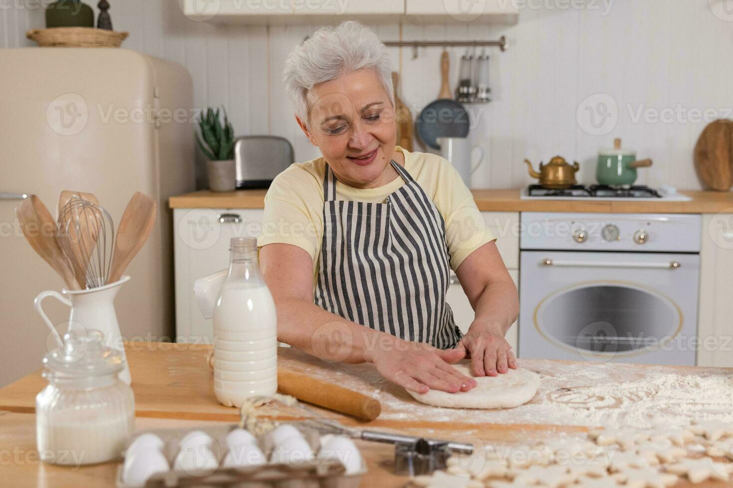 Happy senior woman cooking in kitchen. Stylish older mature gray haired lady grandmother knead dough bake cookies. Old grandma cook homemade food. Household housewife housework concept. photo