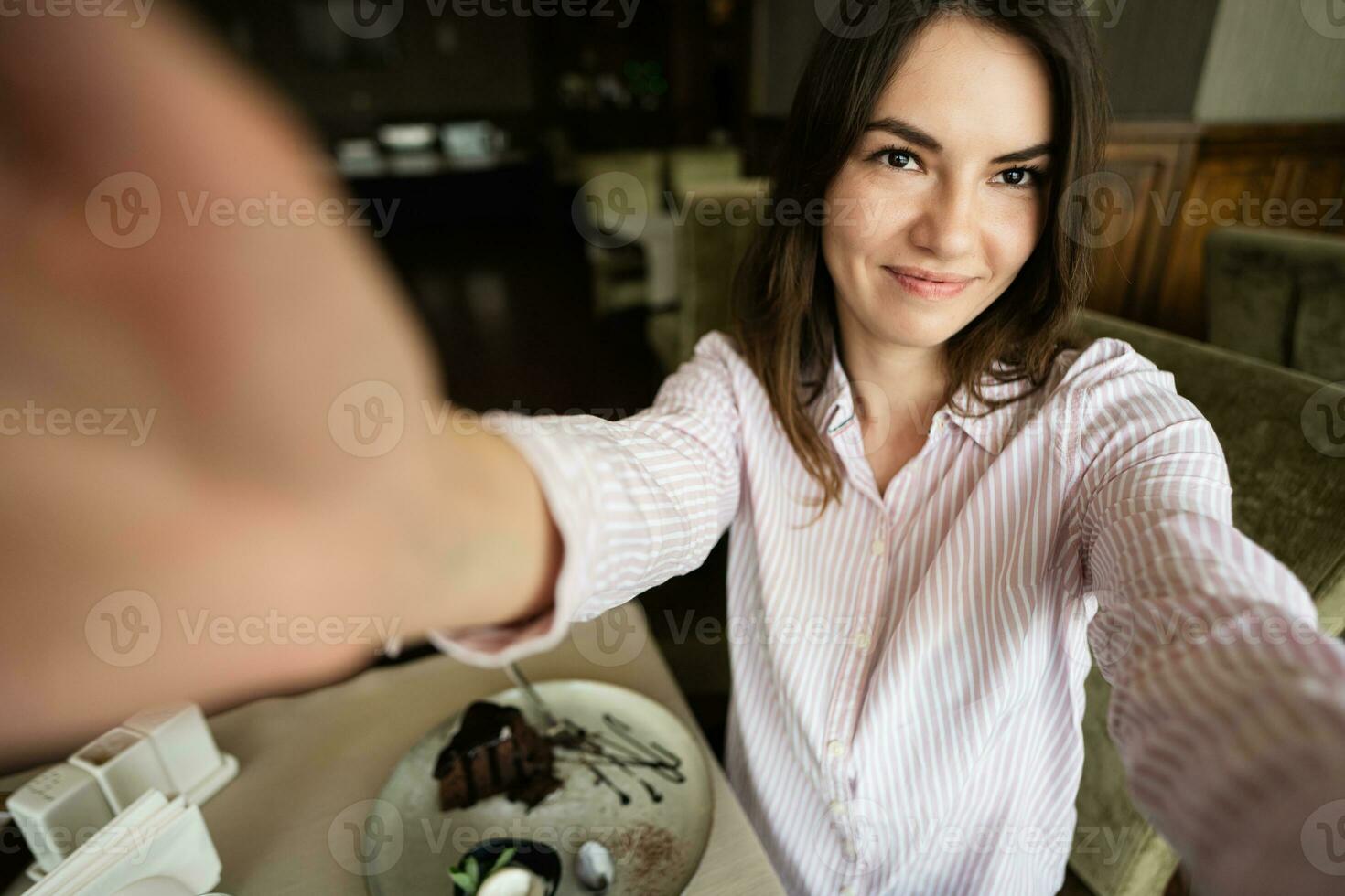 Young beautiful brunette woman sit in coffee shop cafe restaurant indoors, hold camera and making selfie. photo
