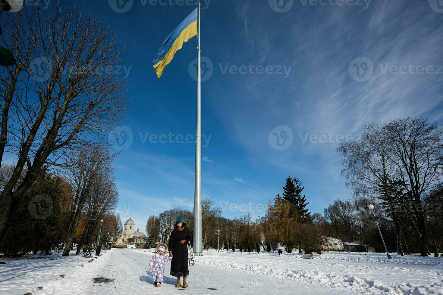 Mother and child walking on a sunny frosty winter day in the park, against background of a flagpole with the Ukrainian flag. photo
