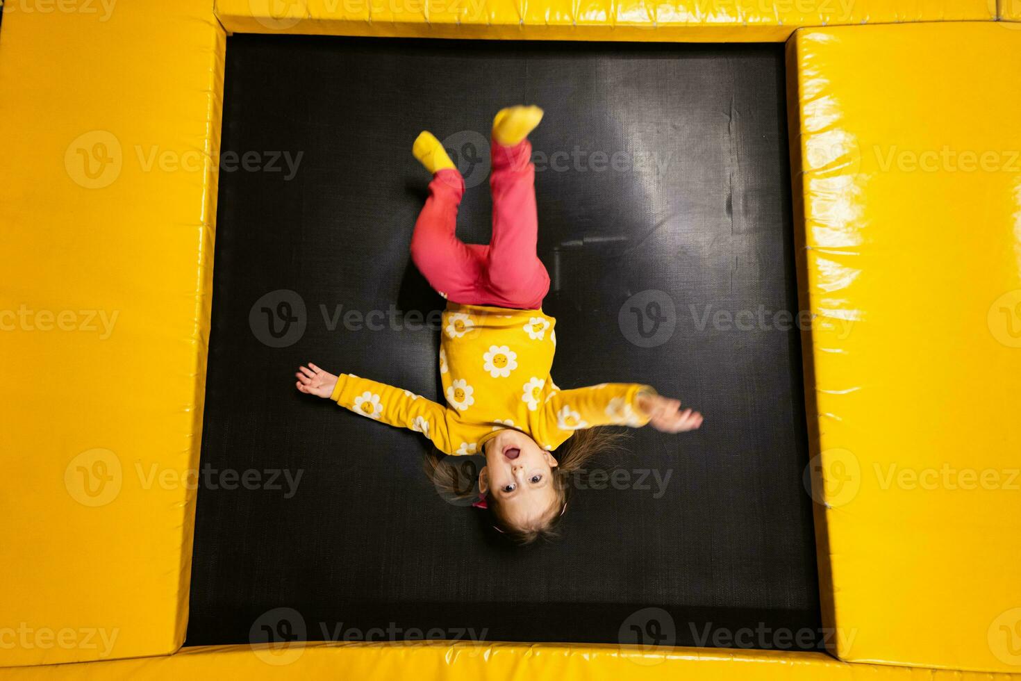 Baby girl kid lies upside down on trampoline at playground park. Child in motion during active entertaiments. photo