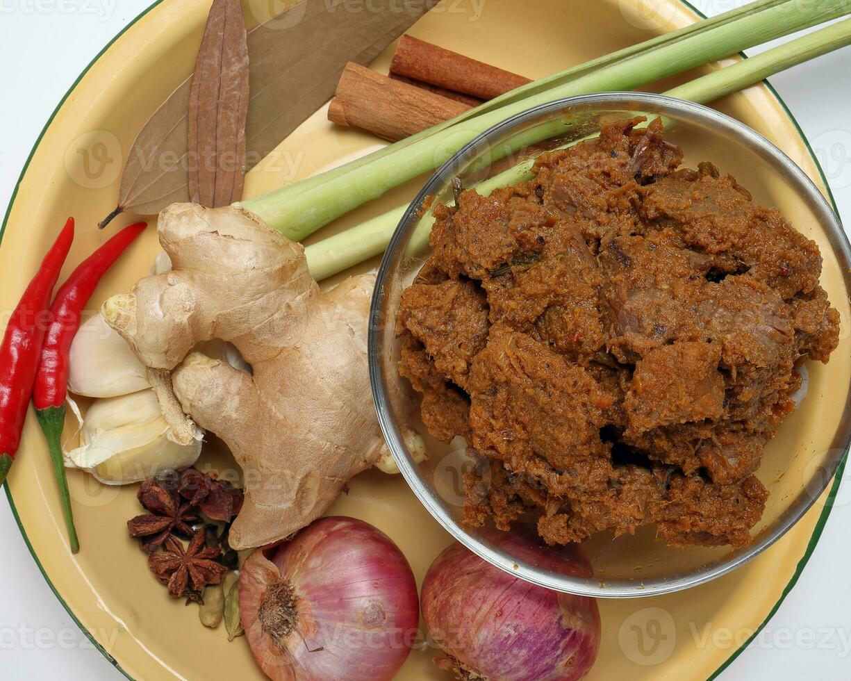 Traditional cooked beef meat rendang dry curry closeup in transparent glass bowl with ingredients onion ginger lemon grass bay leaf cardamom clove cinnamon garlic chilli star anise photo