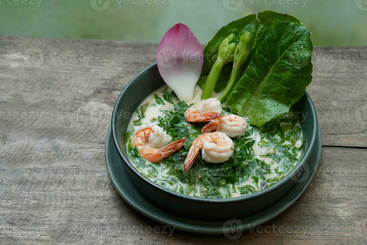 closeup four shrimp based coconut soup with gnetum gnemon leaves and prawns on green ceramic bowl on green ceramic plate, on wooden table, blur nature background, food, nature, copy space photo