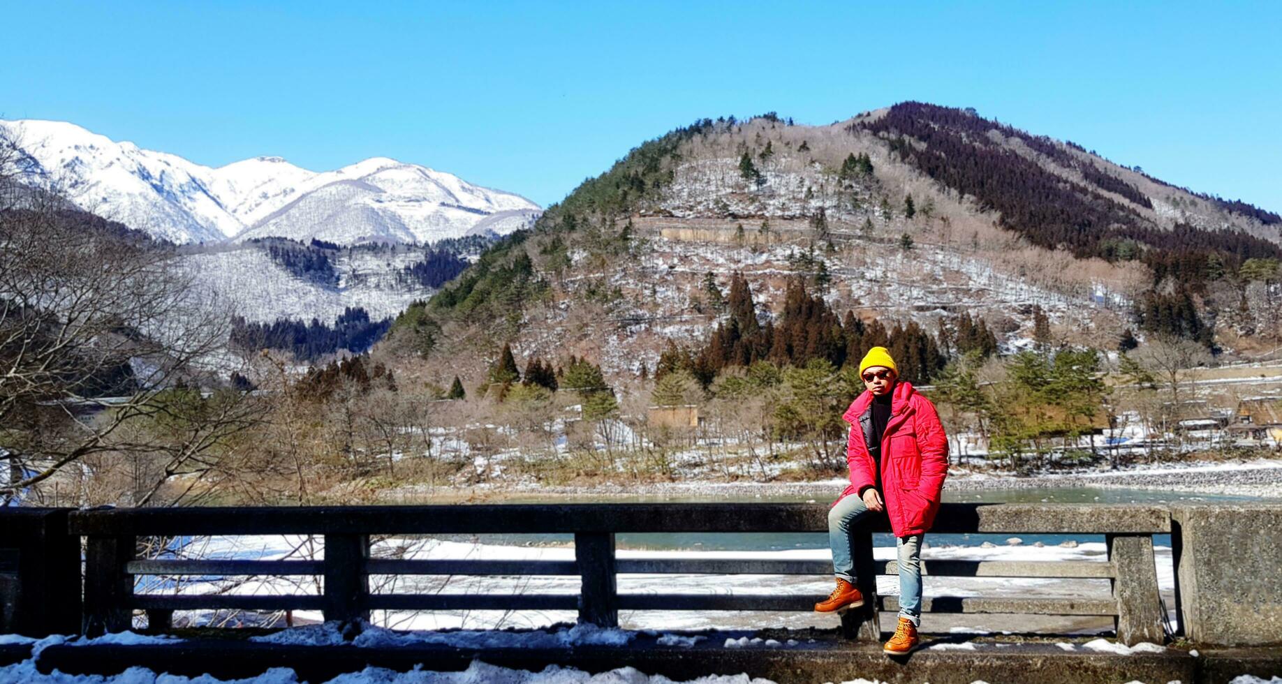 Tourist man in red coat and yellow knit hat sits on bridge rail with tree, snow on mountain and clear blue sky background with copy space at Japan. People travel in Asia with natural and landmarks. photo