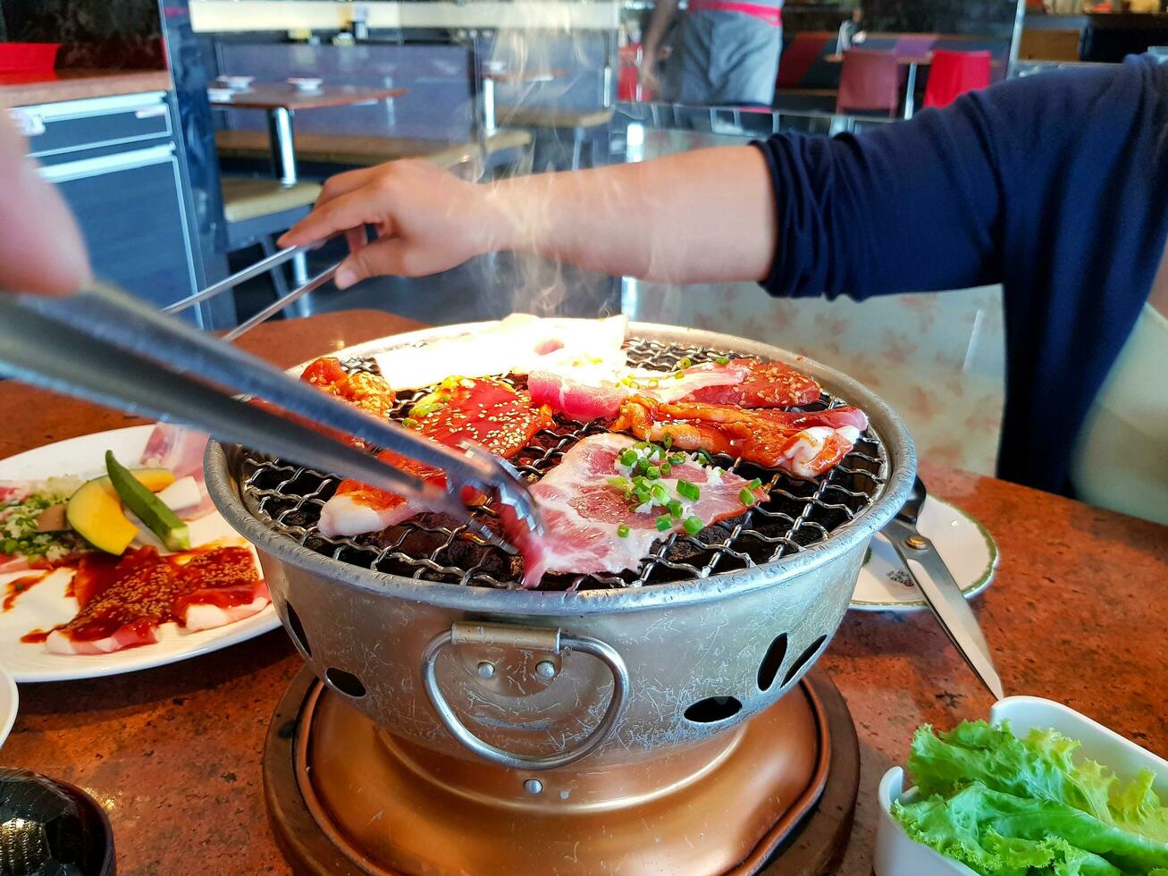 Asian woman grilled raw sliced beef and pork on the hot stainless steel stove with smoke. Barbecue or Lunch time and Japanese style yakiniku. Selective focus on food. photo