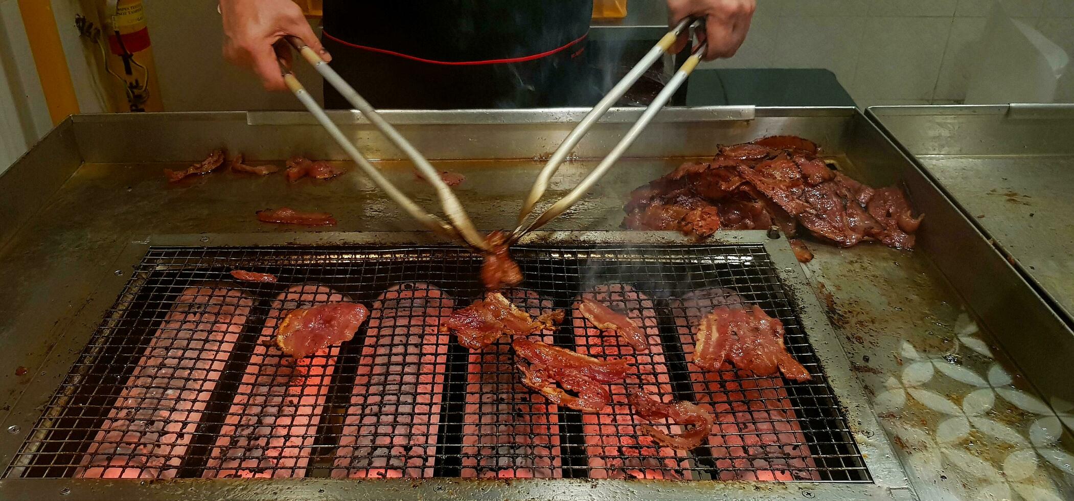 Close up many grilled red pork or barbecue food on the net stove with movement of chef's hand using to cooking utensil in the kitchen at Singapore. Selected focus and Delicious concept photo