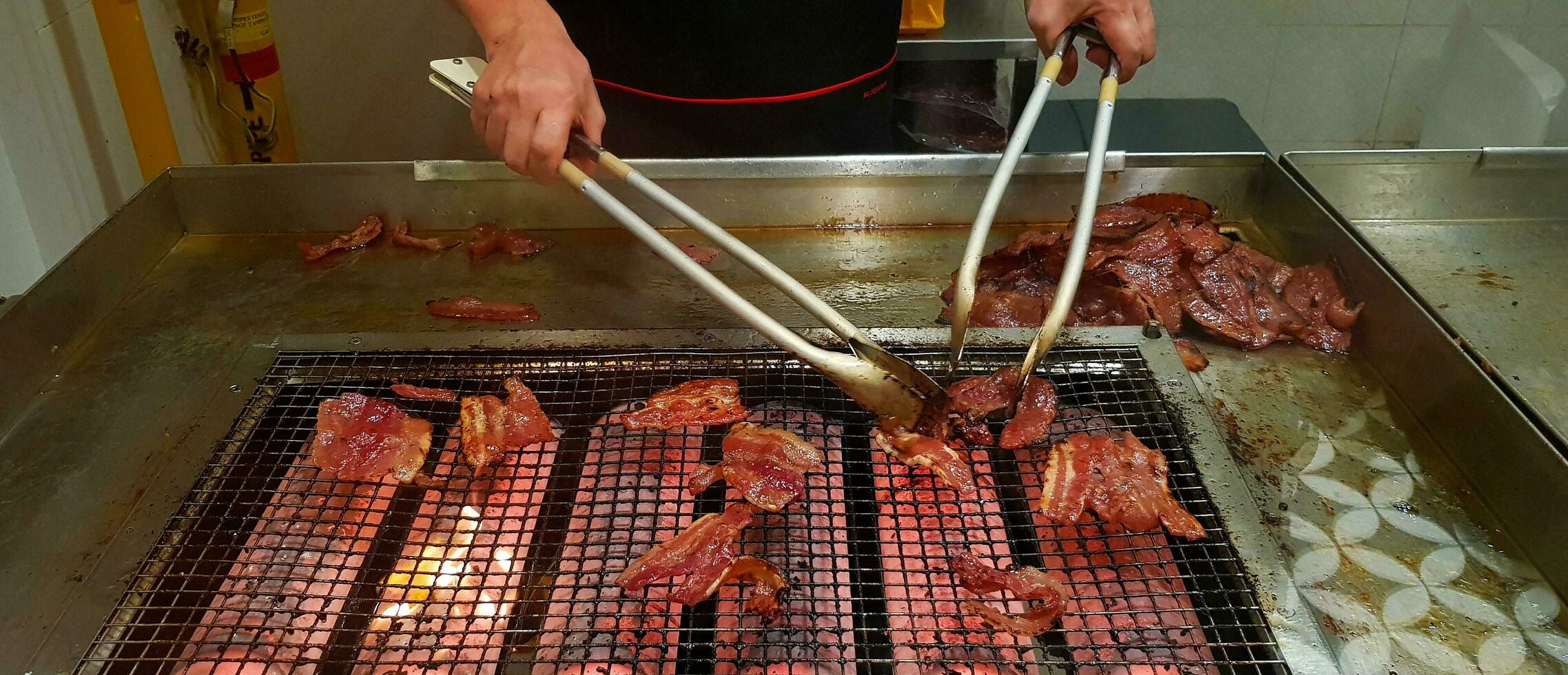 Close up many grilled red pork or barbecue food on the net stove with movement of chef's hand using to cooking utensil in the kitchen at Singapore. Selected focus and Delicious concept photo