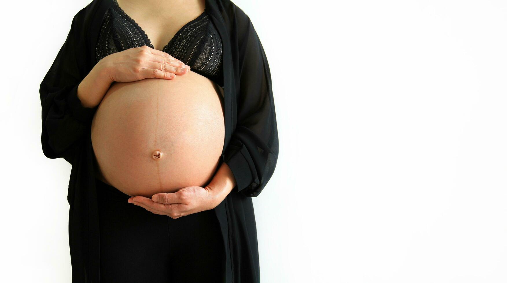 Pregnant woman wearing black dress and holds hands on swollen belly isolated on white background and copy space with clipping path. Pregnancy 7-9 months, motherhood, love, expectation and care baby. photo